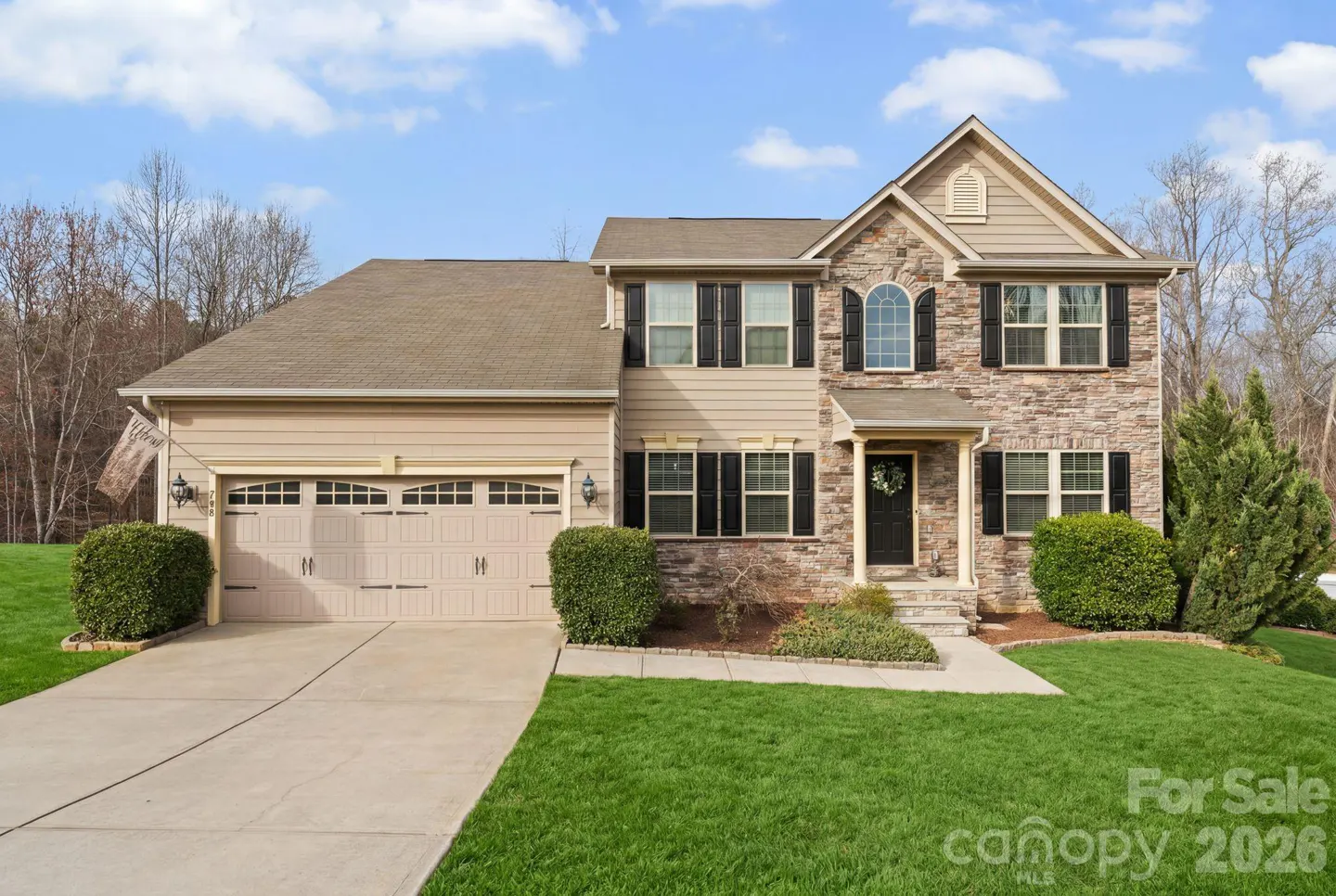 Two-story house with tan siding and stone facade, black shutters, and a two-car garage on a green lawn.