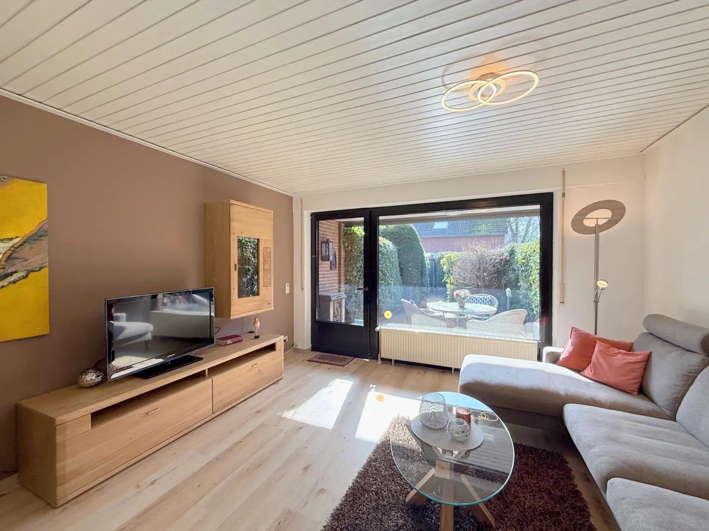Living room with a gray sectional sofa, glass coffee table, and light wood TV stand. A large window overlooks a patio.