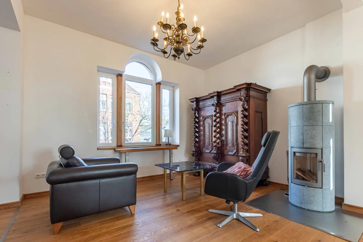 Living room with wood floors, white walls, and a chandelier. There is a fireplace, a dark wood armoire, and two chairs. A window looks out onto a street.