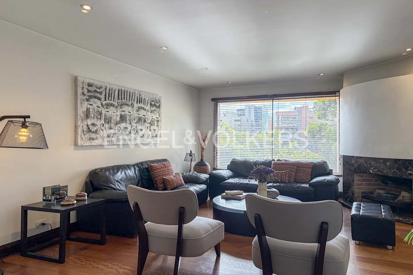 Modern living room with black leather sofas, two light gray armchairs, a round coffee table, and a large abstract painting.