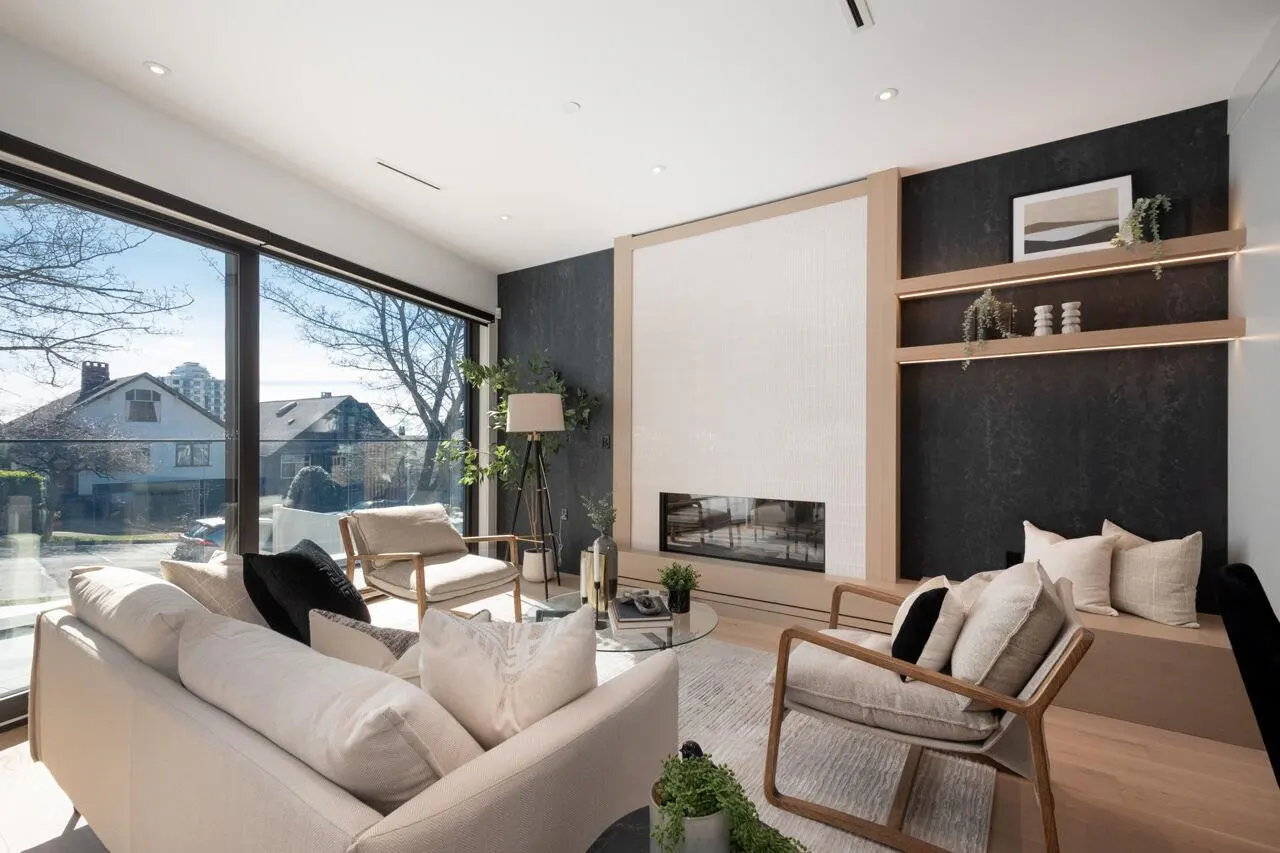 Bright living room with a white sofa, beige chairs, and a modern fireplace. Large windows offer a view of a residential street.