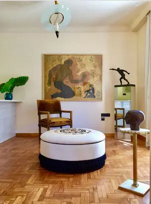 A living room with parquet floors, a white ottoman, a brown chair, and a painting of a man on the wall.
