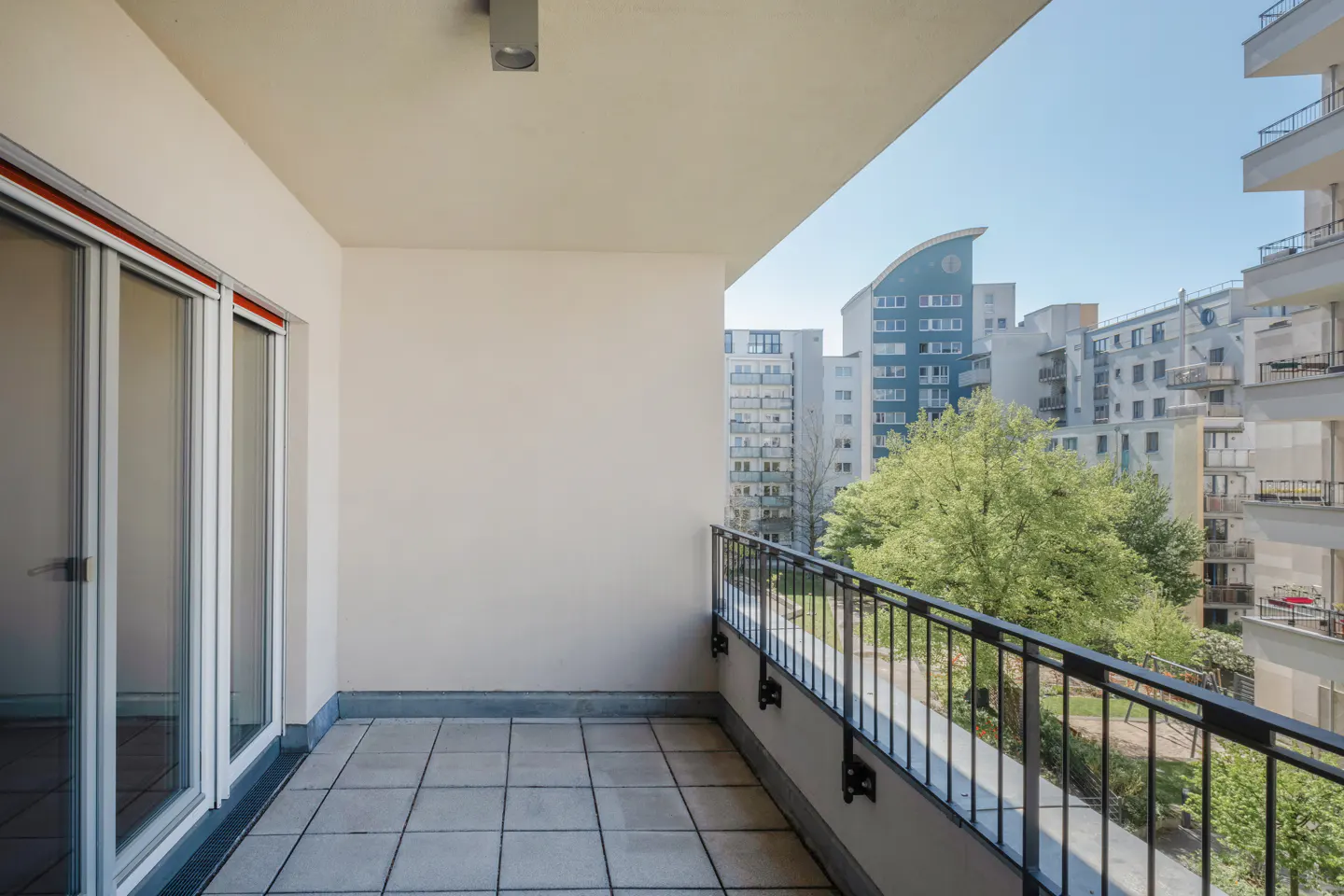 Apartment balcony with tile floor, black metal railing, and sliding glass doors. City buildings and green trees are visible in the background.