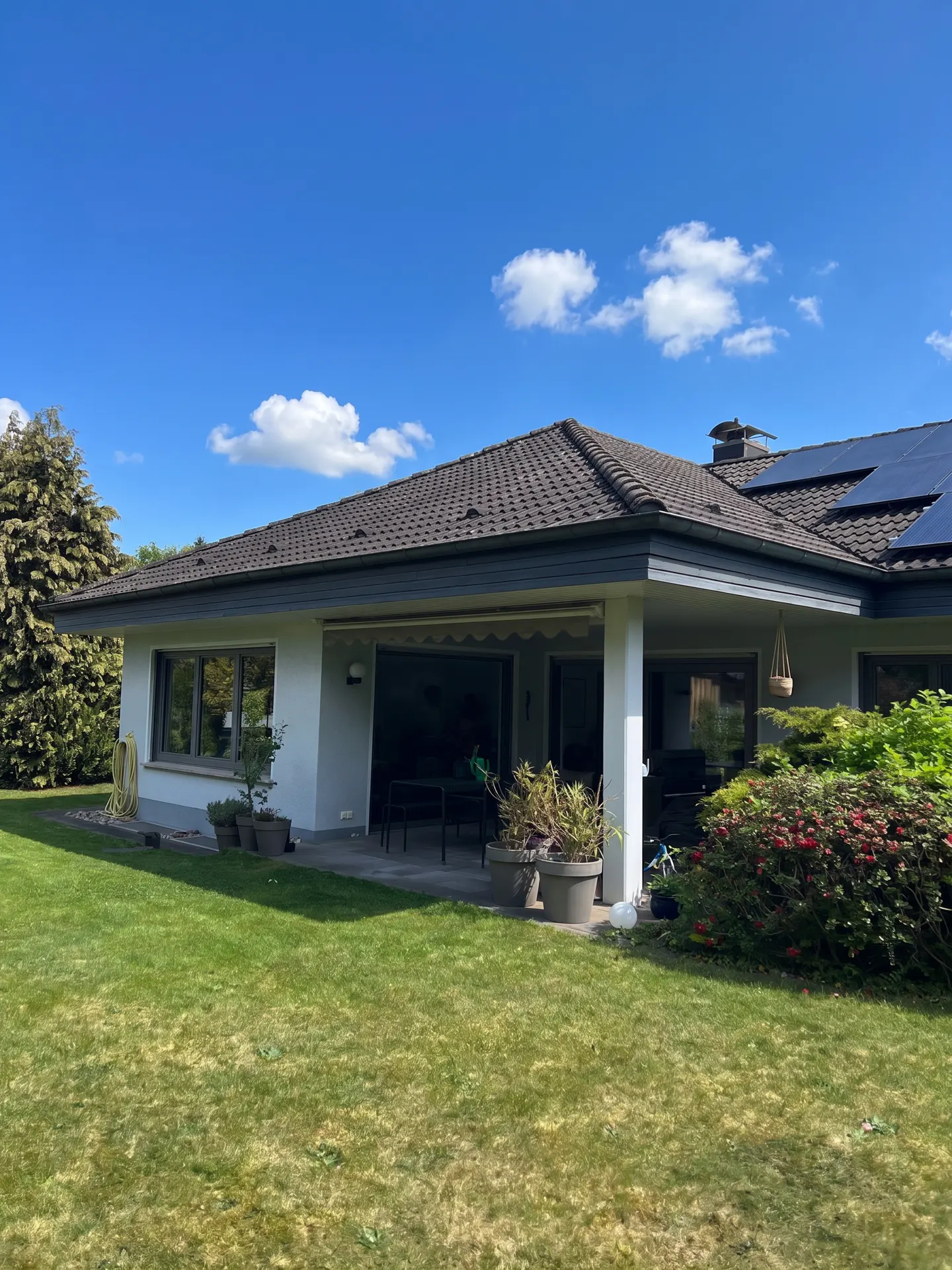 Backyard view of a white house with a dark tiled roof, solar panels, a covered patio, and a lush green lawn.
