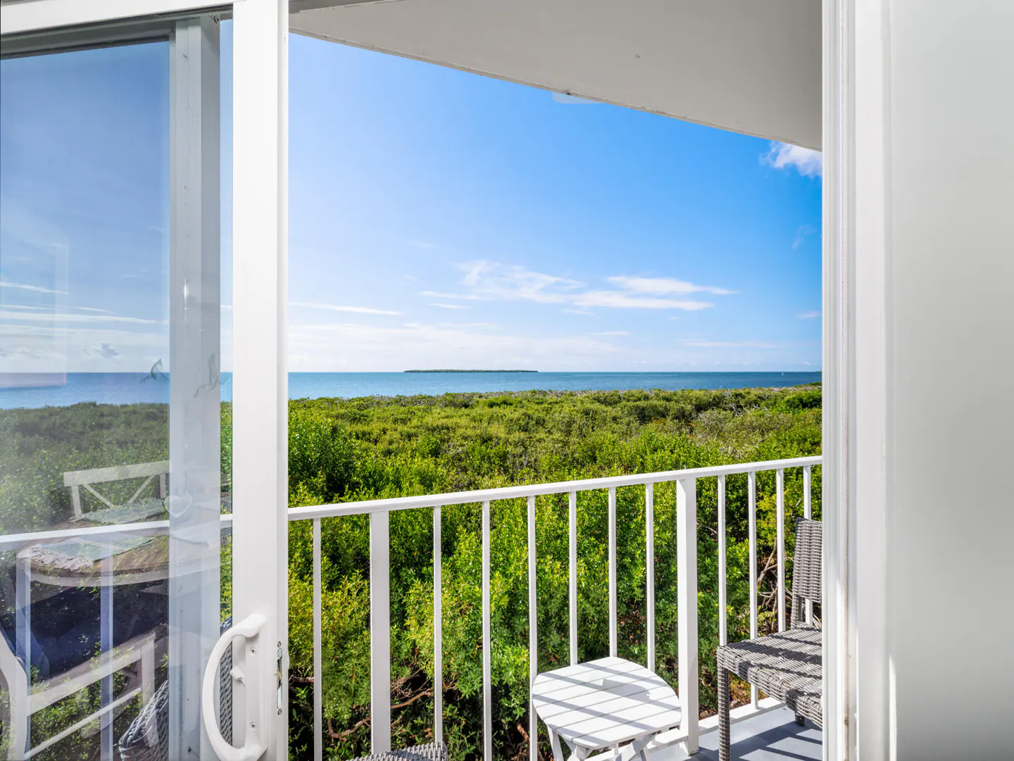 Balcony view with white railing, table, and chair overlooking lush green trees and the ocean under a clear blue sky.