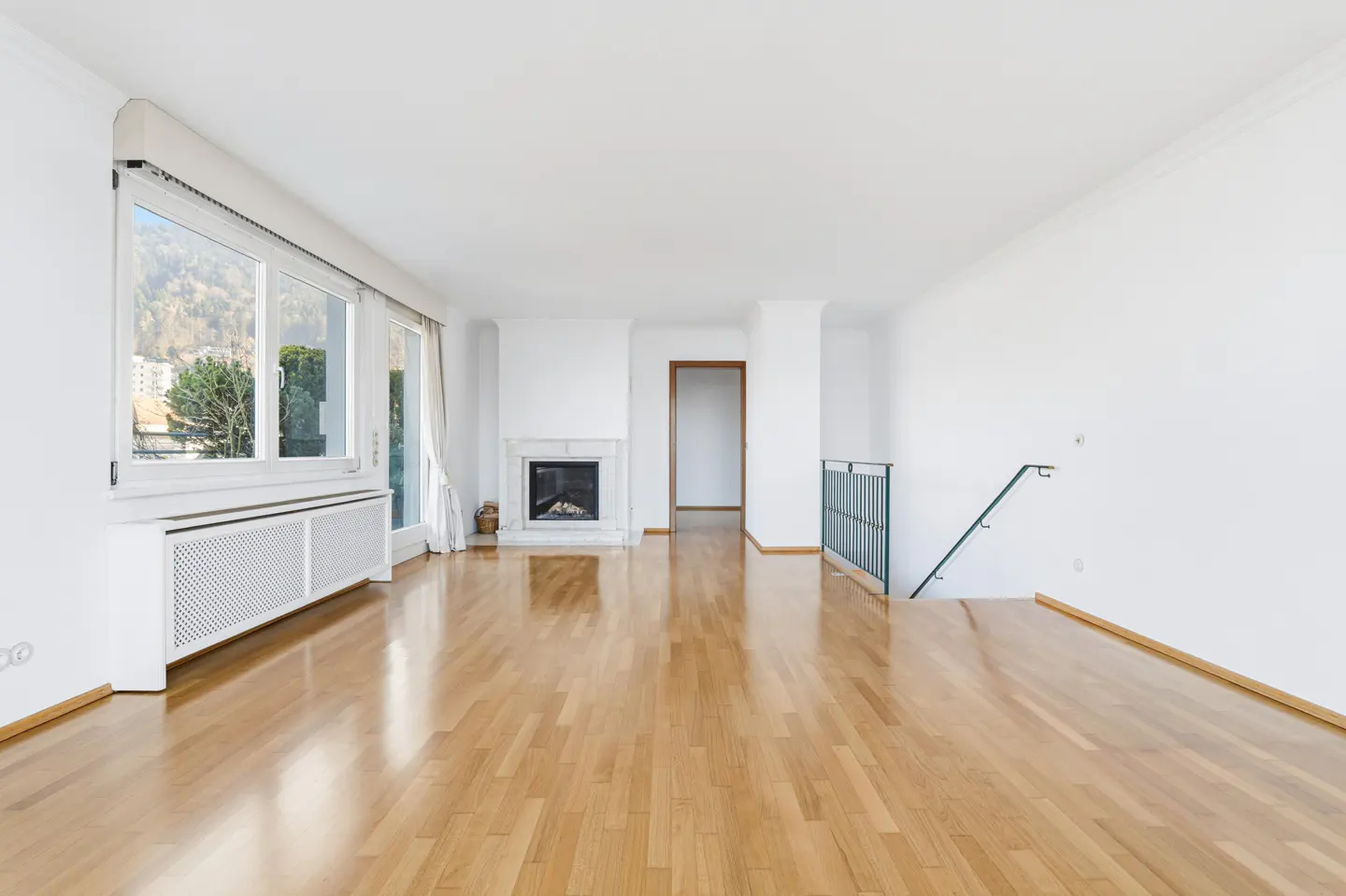 Bright, empty living room with hardwood floors, white walls, fireplace, large windows, and a staircase with a black railing.