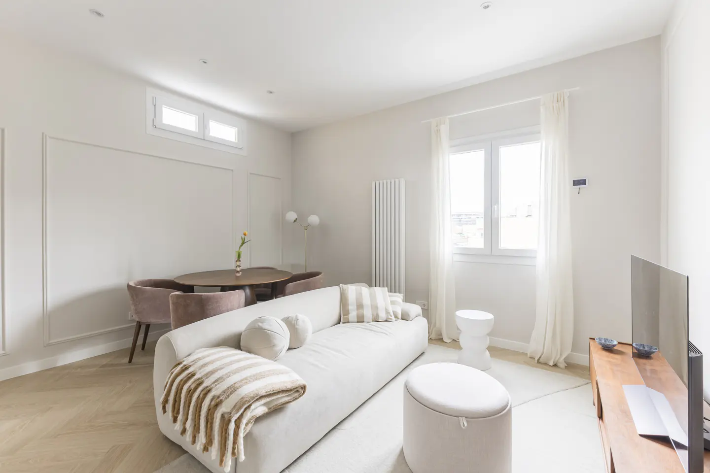 Bright living room with white sofa, round wood table with pink chairs, and a TV on a wood stand.