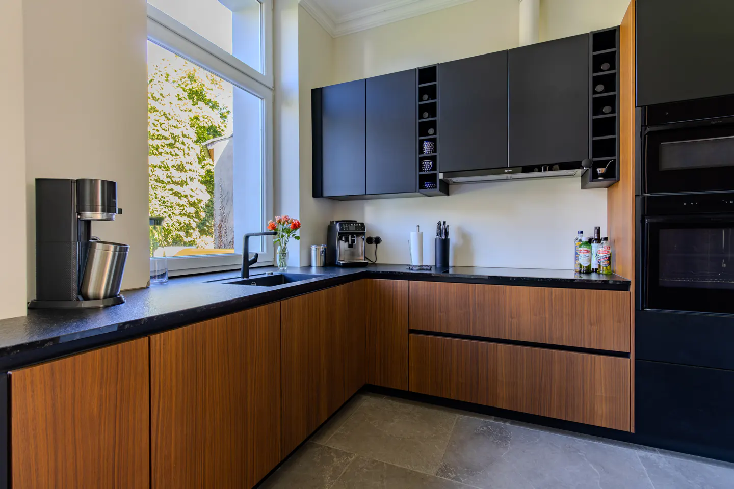 Modern L-shaped kitchen with wood-grain lower cabinets, matte black upper cabinets, black countertops, and a window view.