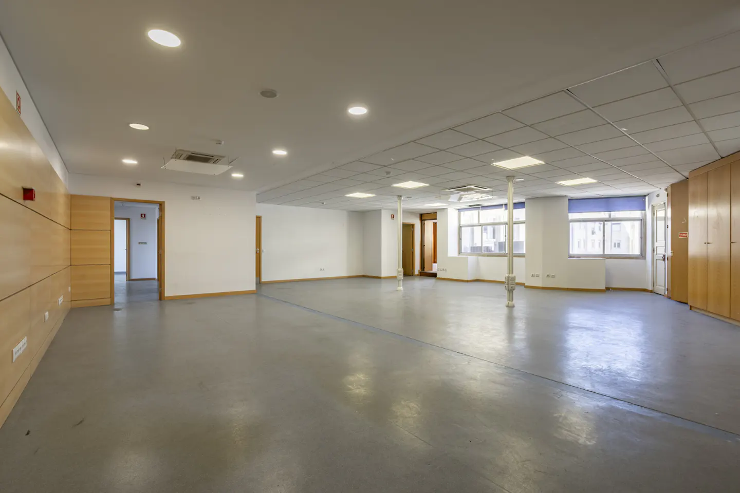Empty office space with gray floors, white walls, and wood accents. Windows with blue shades provide natural light.