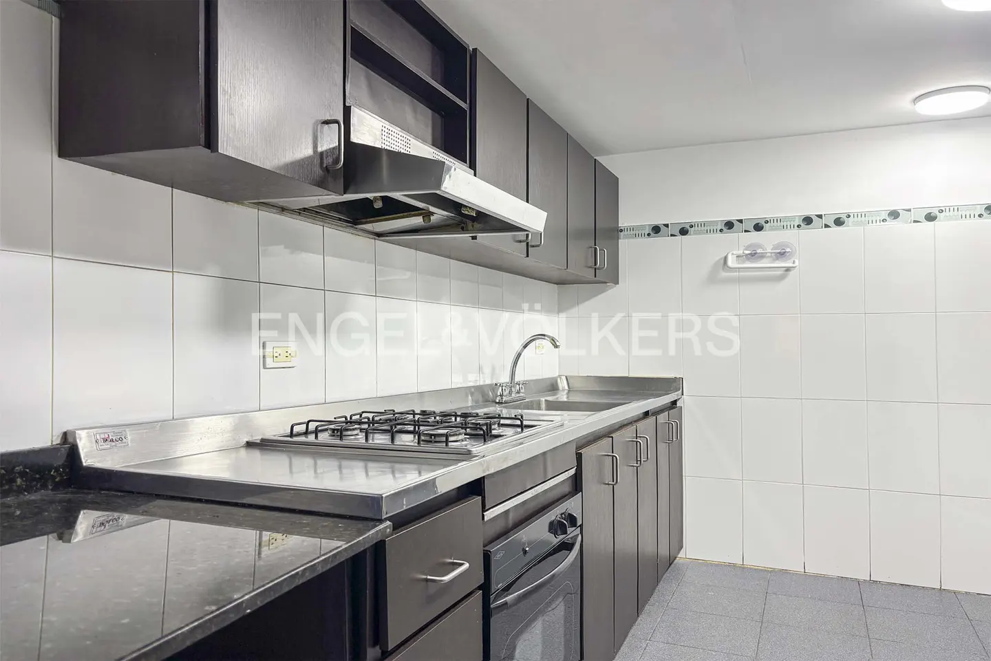 Kitchen with dark brown cabinets, stainless steel gas stovetop, built-in oven, and white tiled walls and floor.