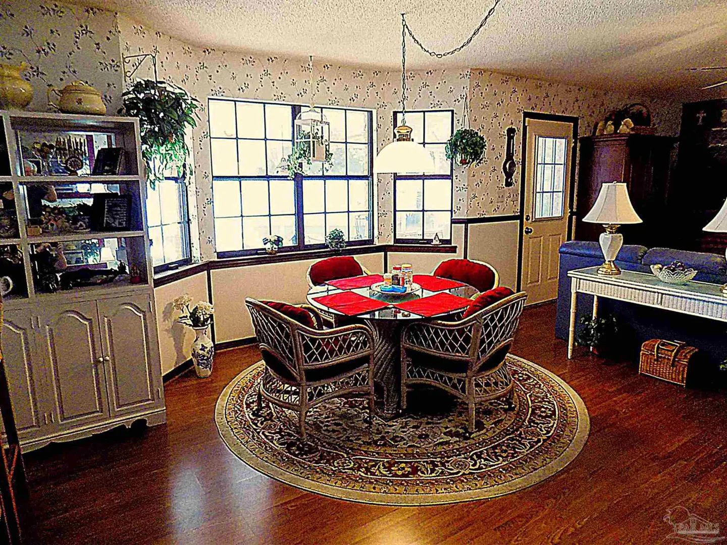 A dining area with a round glass table, four wicker chairs with red cushions, and a patterned rug on a wood floor.
