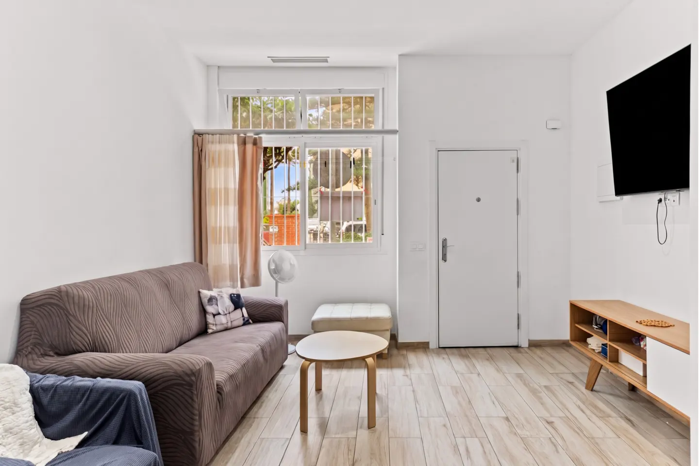 Living room with a brown sofa, round table, and TV. A window with bars and curtains lets in natural light. The walls are white, and the floor is wood.