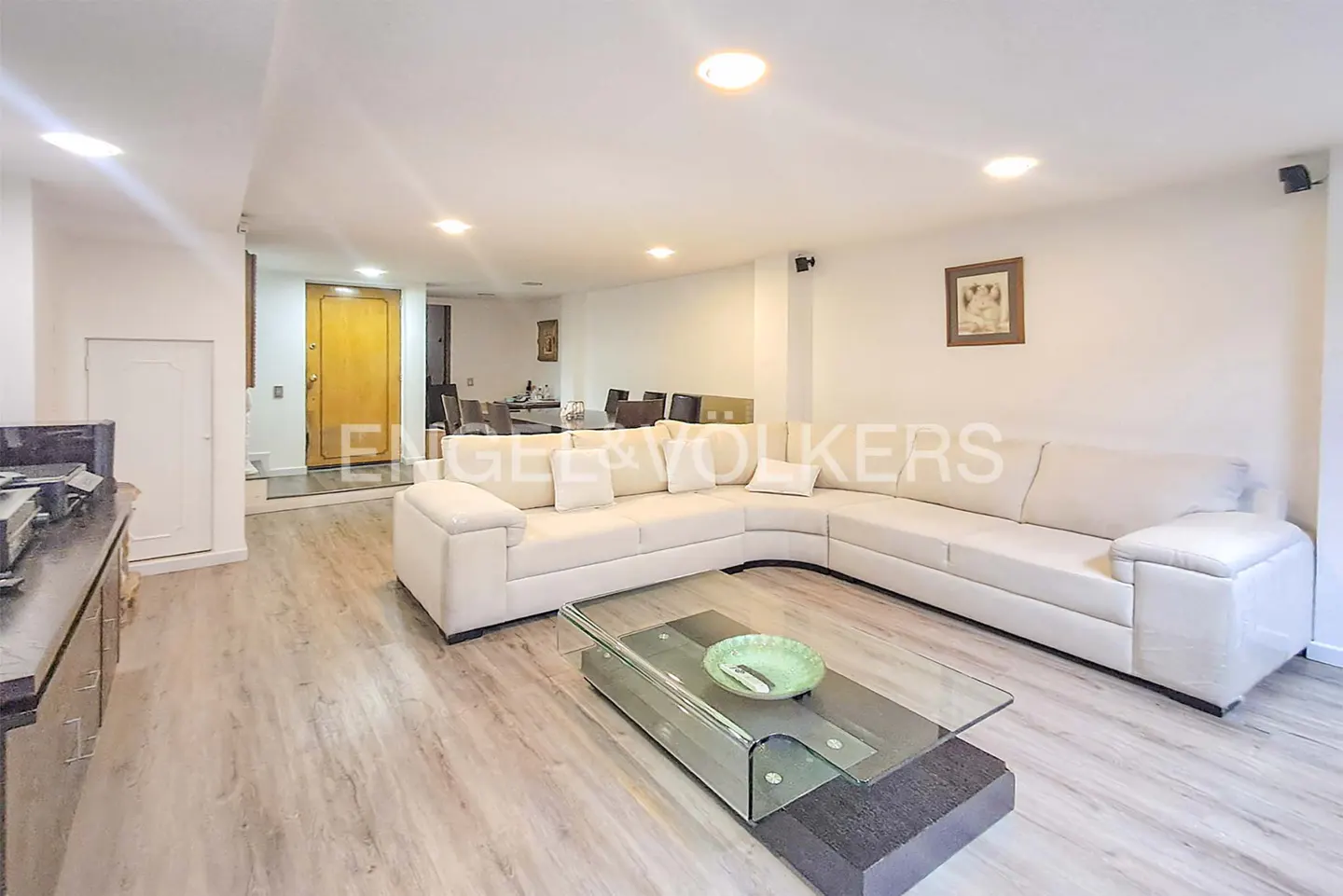A bright living room with a large white sectional sofa, glass coffee table, and light wood floors.