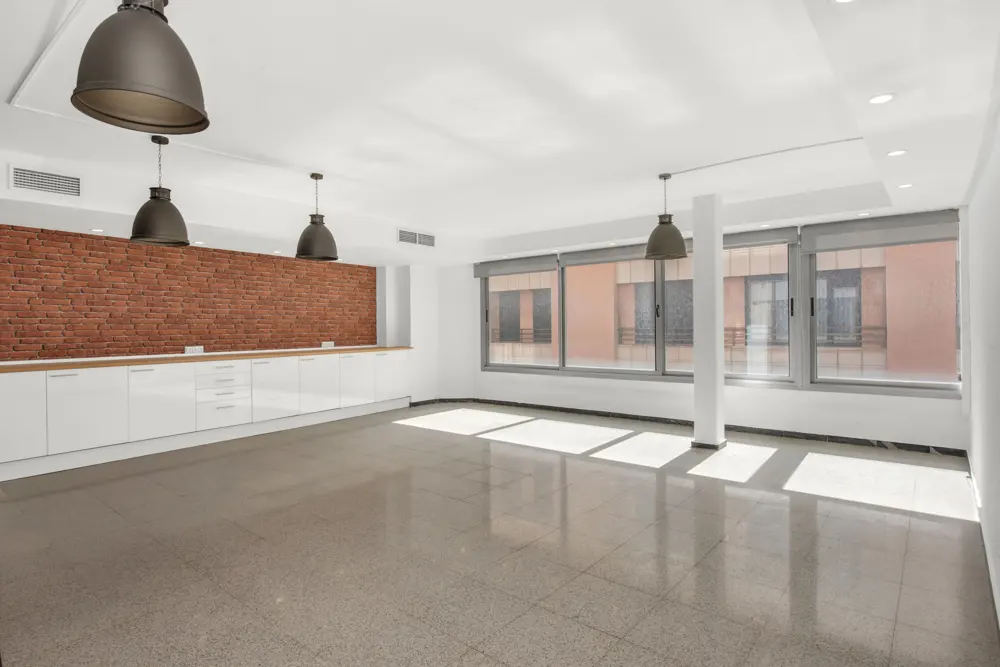 Bright, empty room with white walls, gray tile floor, and large windows. A brick wall and white cabinets line one side, with pendant lights hanging above.