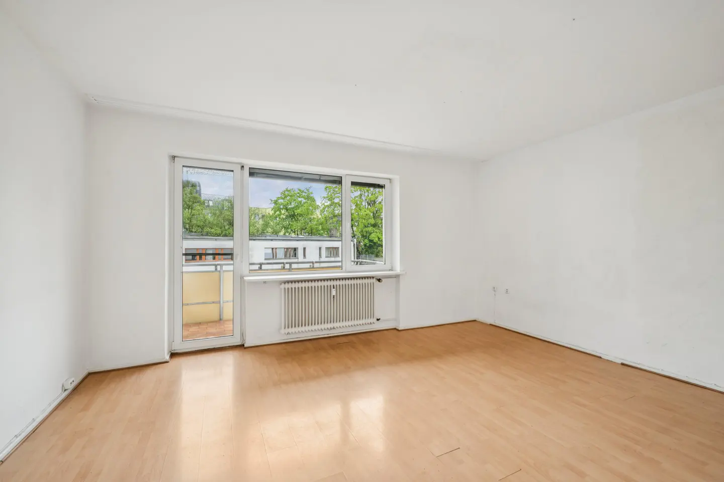 Empty room with light wood floors, white walls, and a window to a balcony with green trees outside.