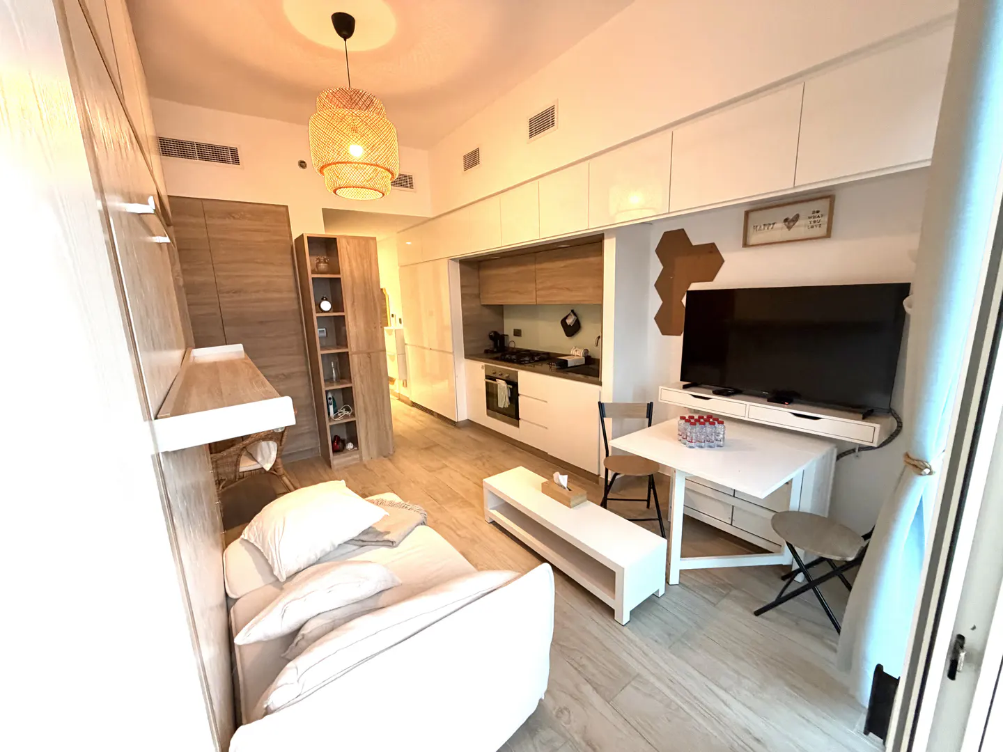 A bright studio apartment with a white sofa, a white table, and a kitchen in the background. A woven lamp hangs from the ceiling.