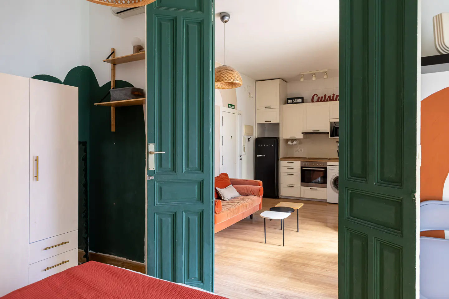 View through green doors into a studio apartment with a kitchen, orange sofa, and white cabinets.
