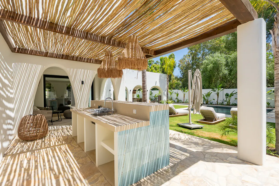 Outdoor kitchen with a sink and blue tile detail, under a thatched roof. Lounge chairs and a pool are visible in the background.