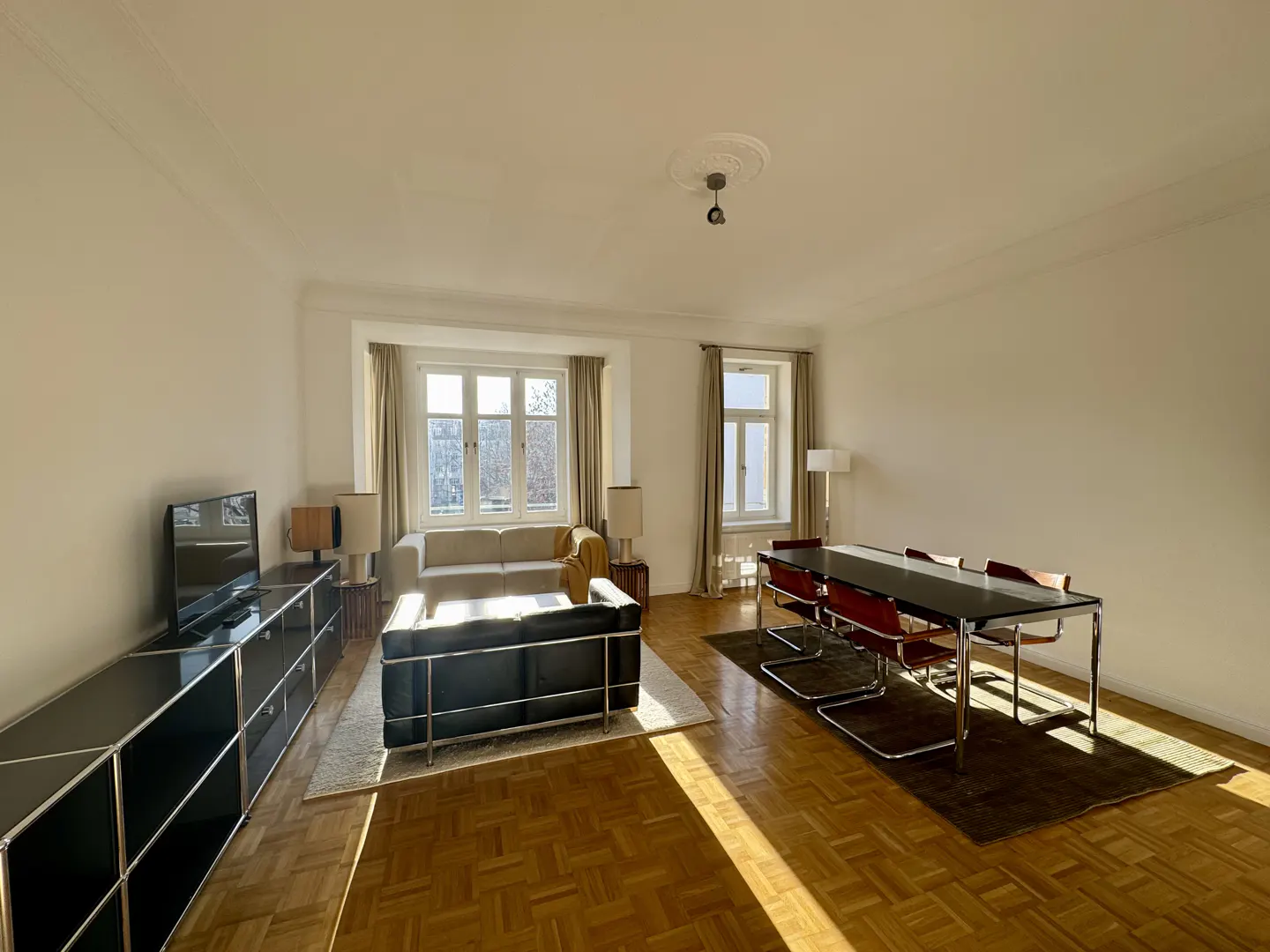 Bright living room with parquet floors, black sofa, beige sofa, black cabinet, and a dining table with red chairs.