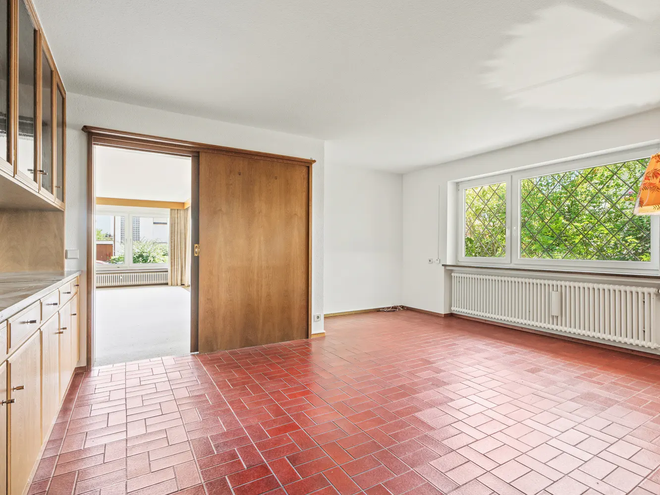 Bright room with red tiled floors, white walls, a large window with a radiator, and a wooden sliding door to another room.