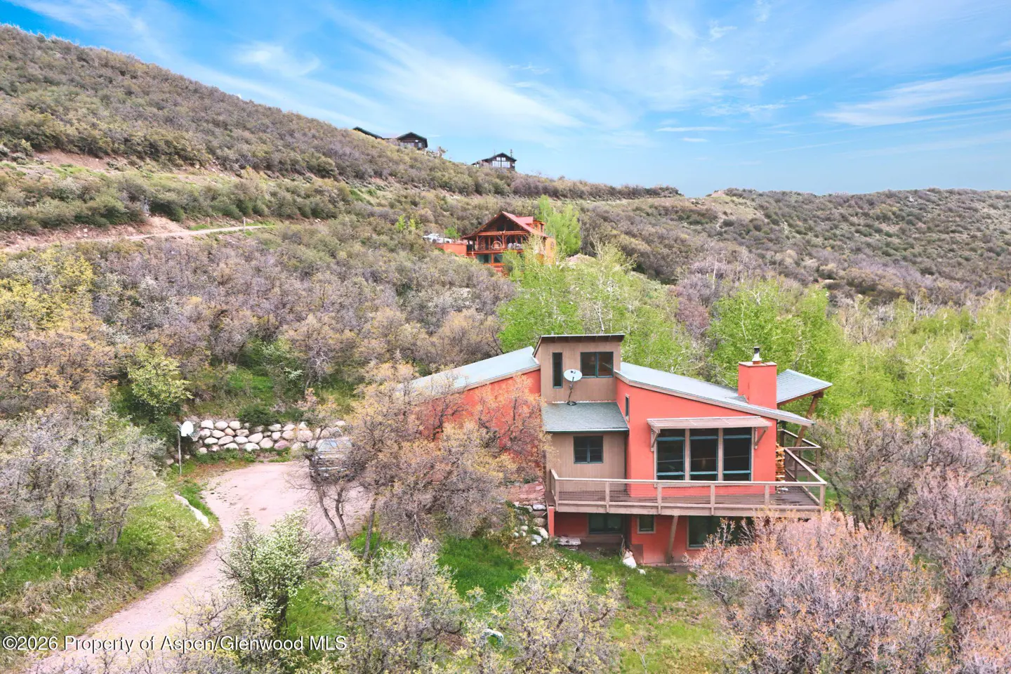 Exterior view of a red house with a deck, nestled on a hillside with trees and a blue sky.