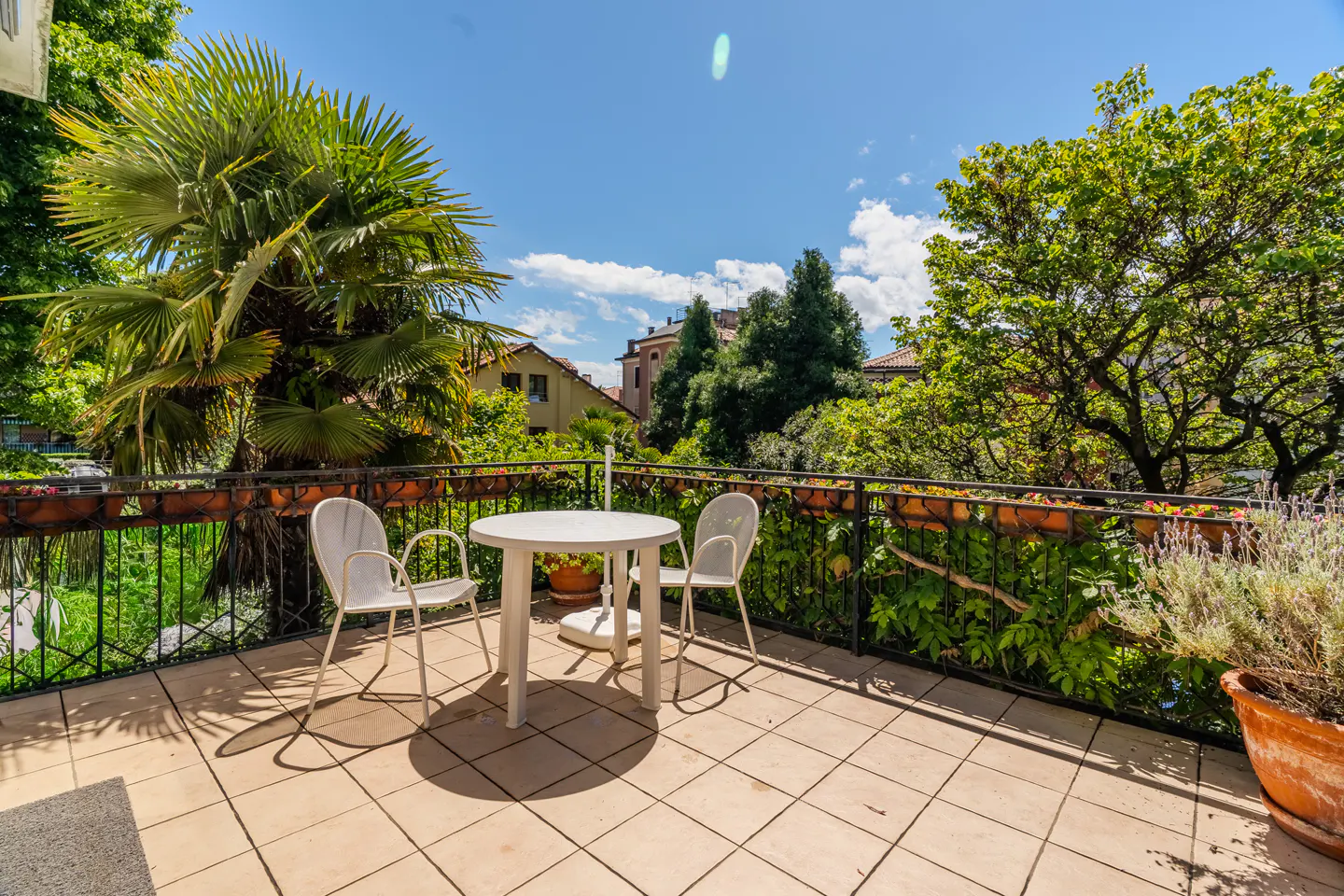 Outdoor patio with a round white table and two white chairs. Lush green trees and plants surround the black metal railing. Blue sky above.