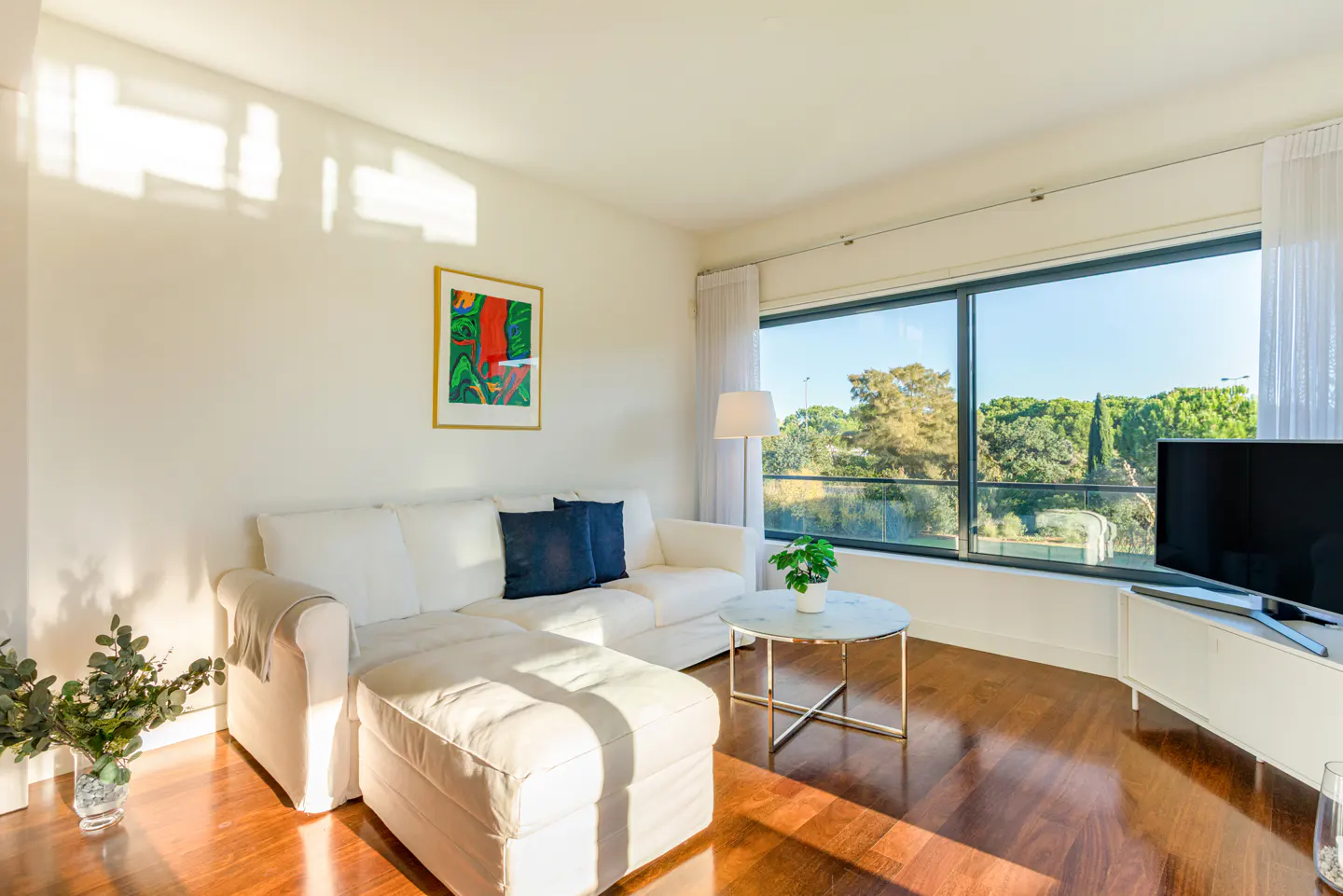 Bright living room with white sofa, blue pillows, and a marble-topped table. Large windows offer a view of trees. Hardwood floors reflect sunlight.