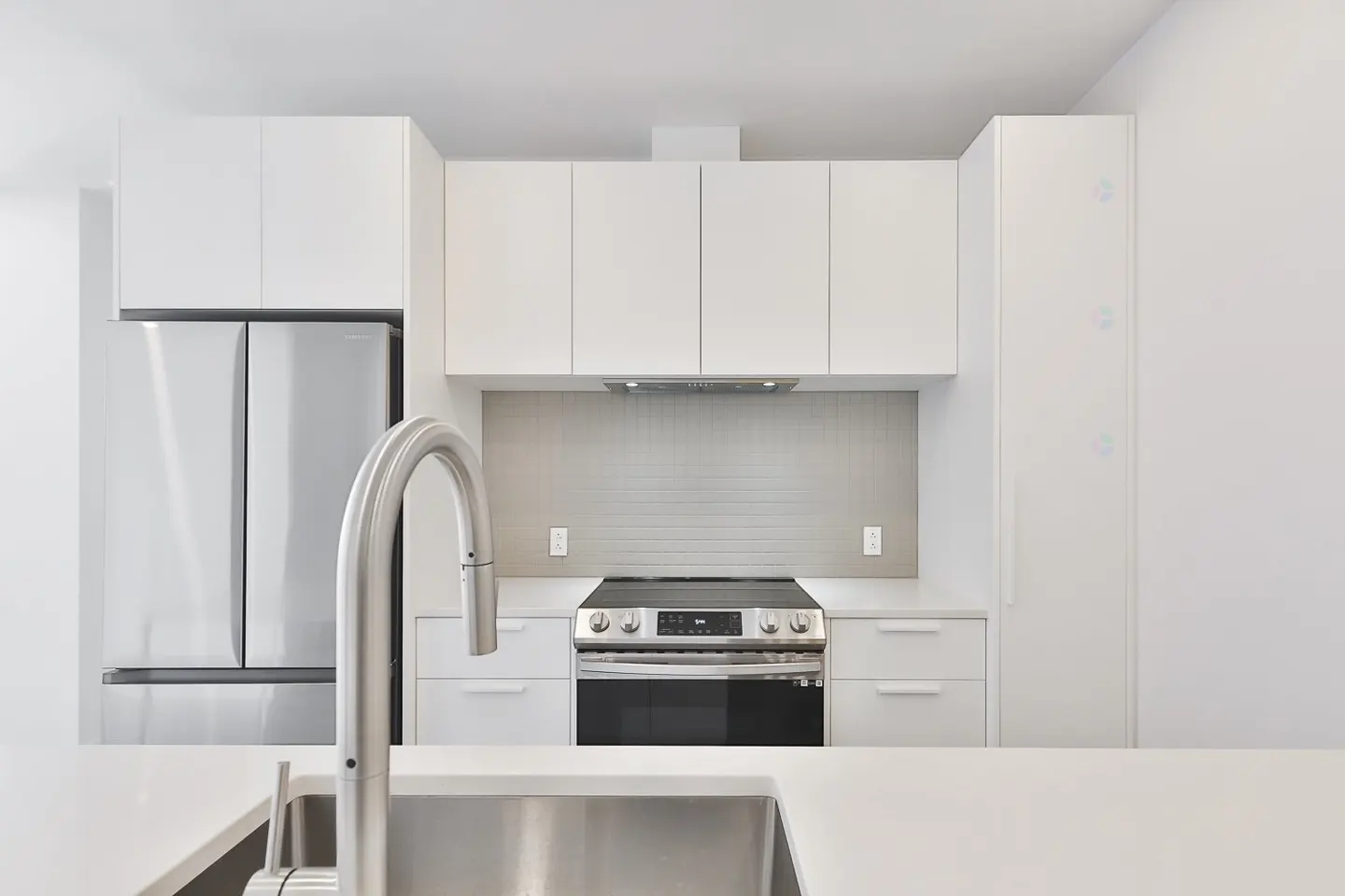Modern kitchen with stainless steel sink, faucet, fridge, and oven. White cabinets and countertops create a clean, minimalist design.