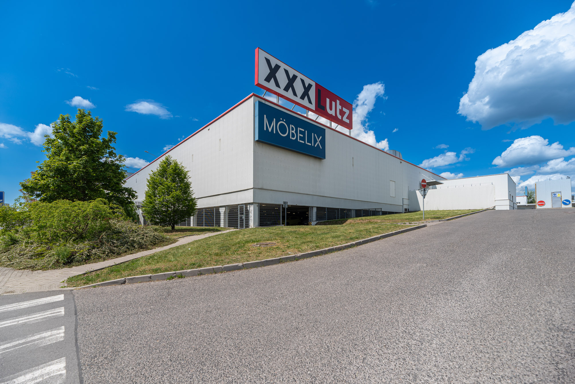 Exterior view of a large, white XXX Lutz Möbelix furniture store under a blue sky with scattered clouds.