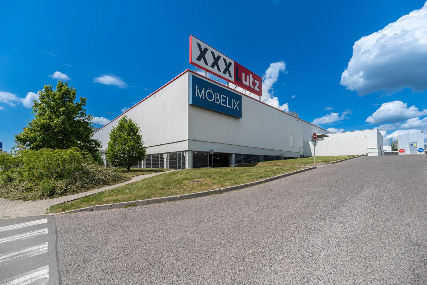 Exterior view of a large, white XXX Lutz Möbelix furniture store under a blue sky with scattered clouds.