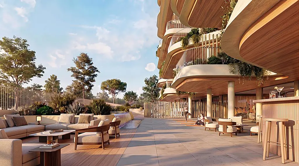 Outdoor patio with beige seating, a bar, and a view of a modern building with curved balconies and greenery. Trees and blue sky in the background.