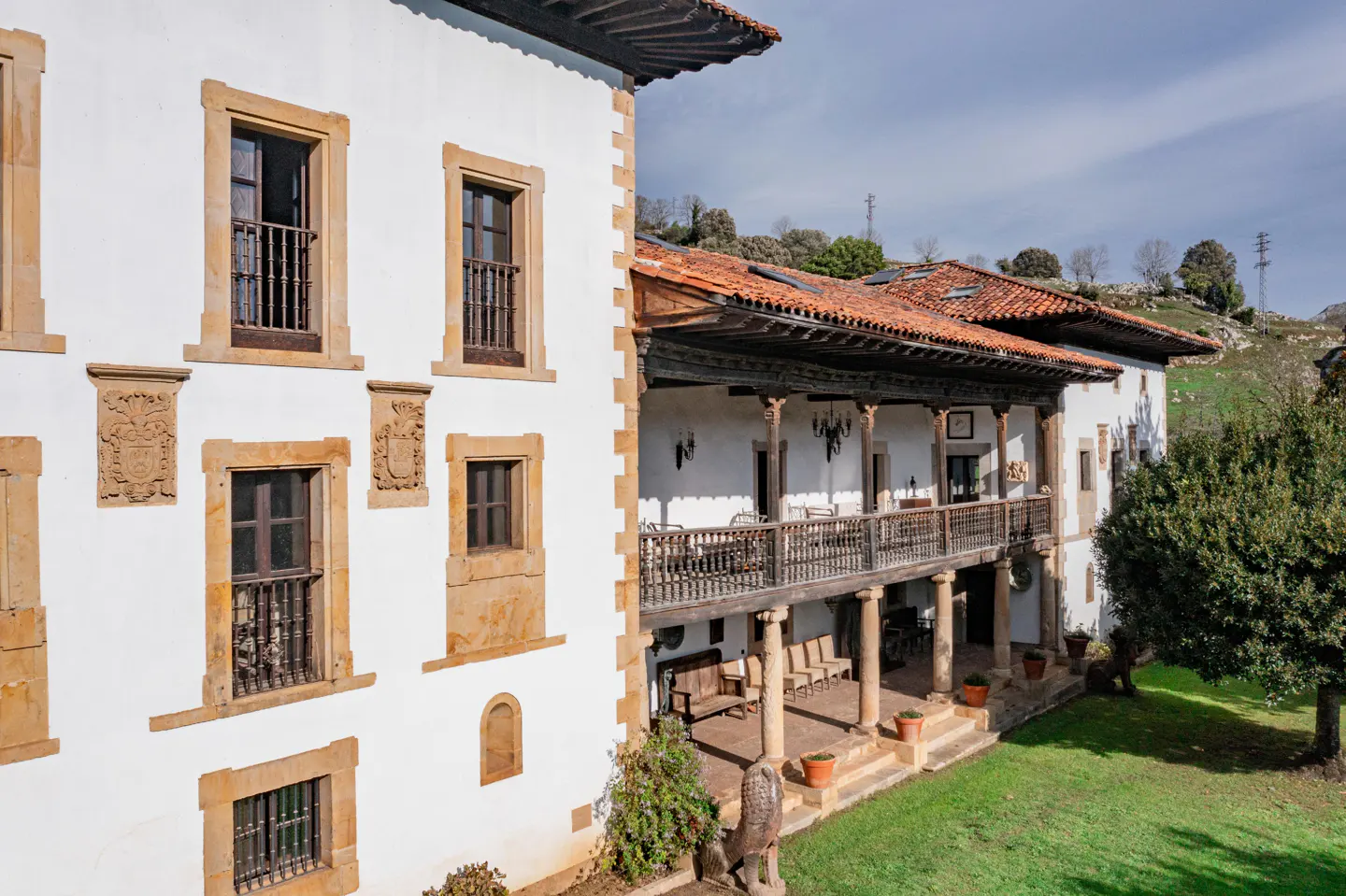 A white historic Spanish manor with stone trim, a wooden balcony, terracotta roof tiles, and a lush green lawn.