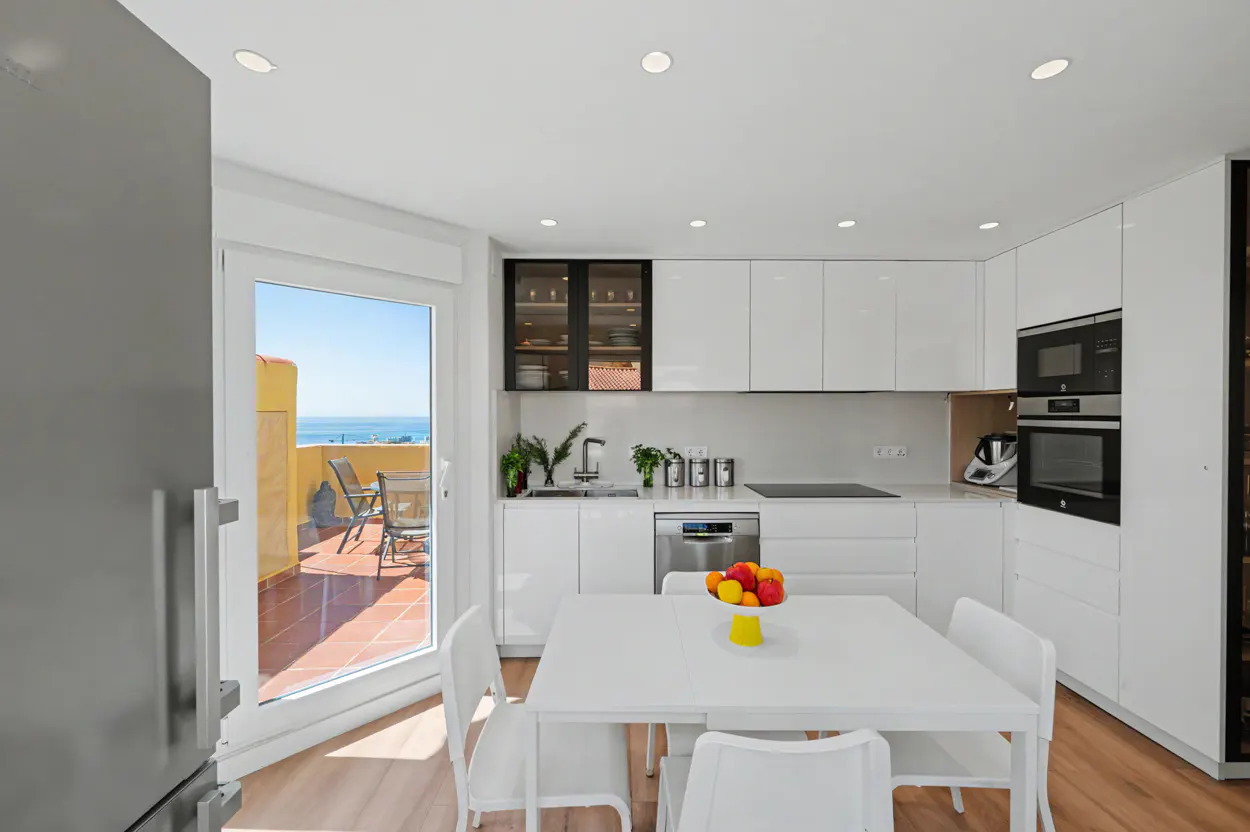 Bright, modern kitchen with white cabinets, stainless steel appliances, and a white table with fruit. Balcony with ocean view visible.