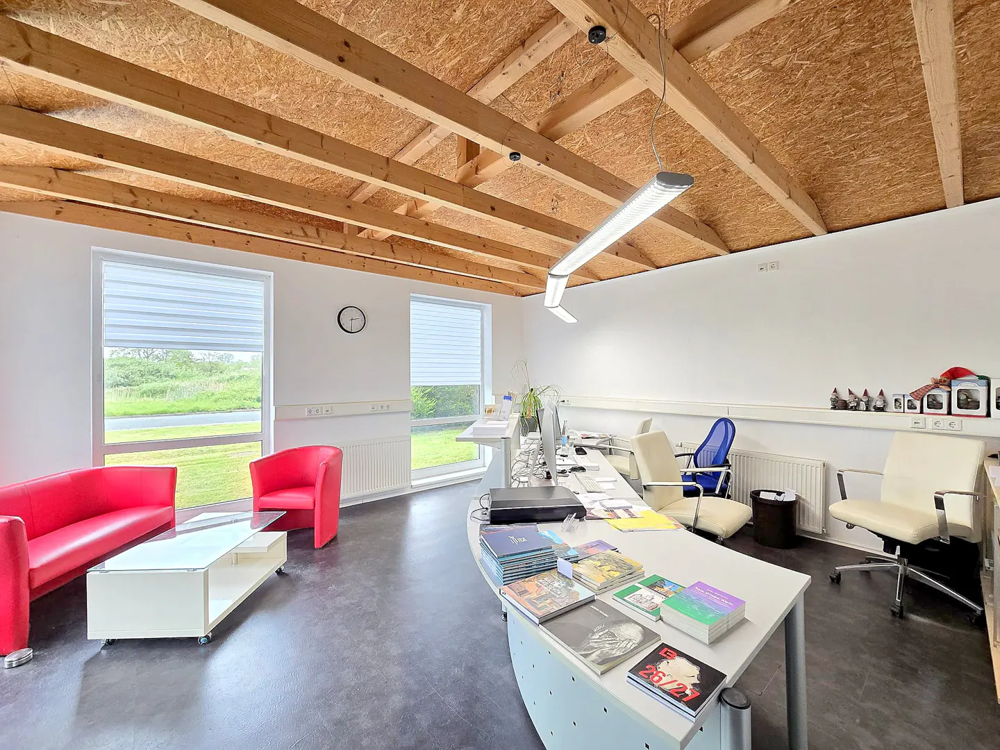 Bright office space with exposed wood beam ceiling, white walls, and gray floor. Red sofa and chair in waiting area, reception desk with books, and office chairs.