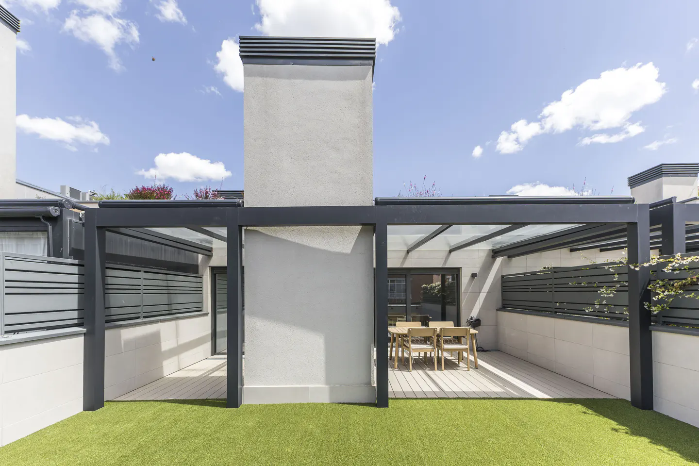 Outdoor patio with a glass-roofed pergola, a dining table with chairs, and artificial grass under a blue sky.
