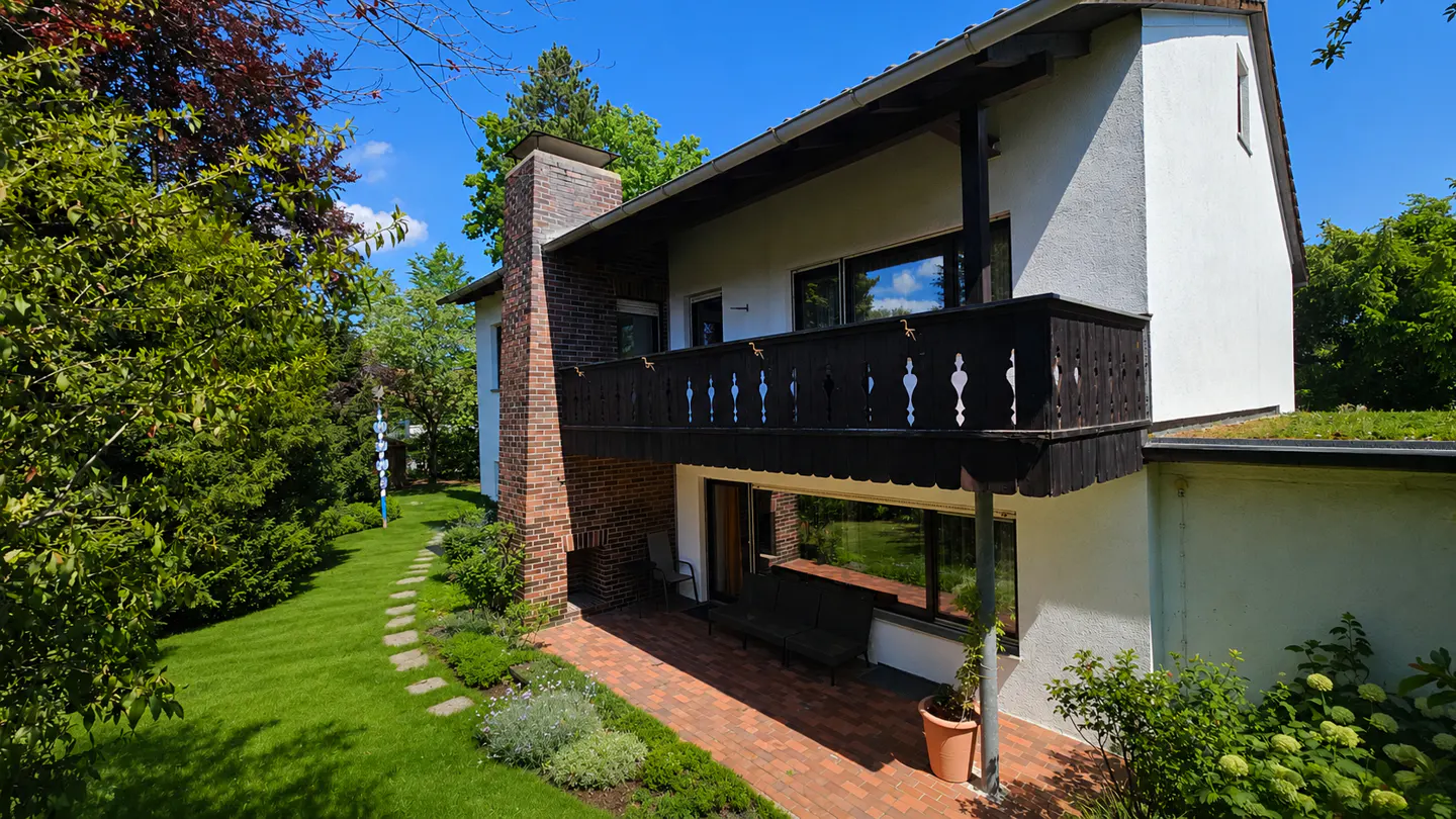 Exterior view of a two-story house with a brick chimney, black balcony, and a brick patio surrounded by green grass and trees.