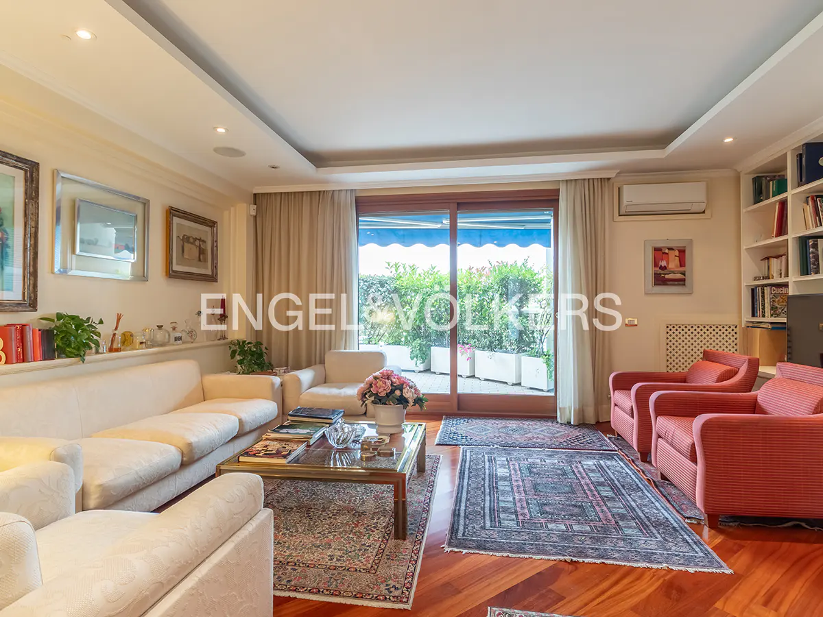 Bright living room with white sofas, red armchairs, and a glass door leading to a balcony with greenery.