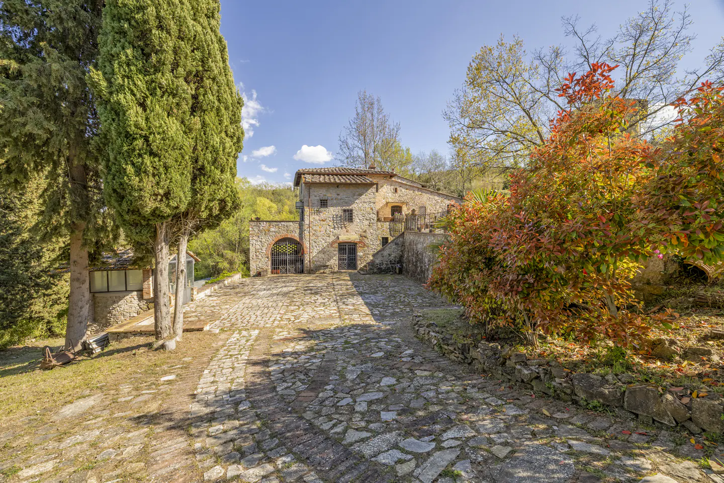 Exterior view of a stone house with a cobblestone driveway, trees, and blue sky.