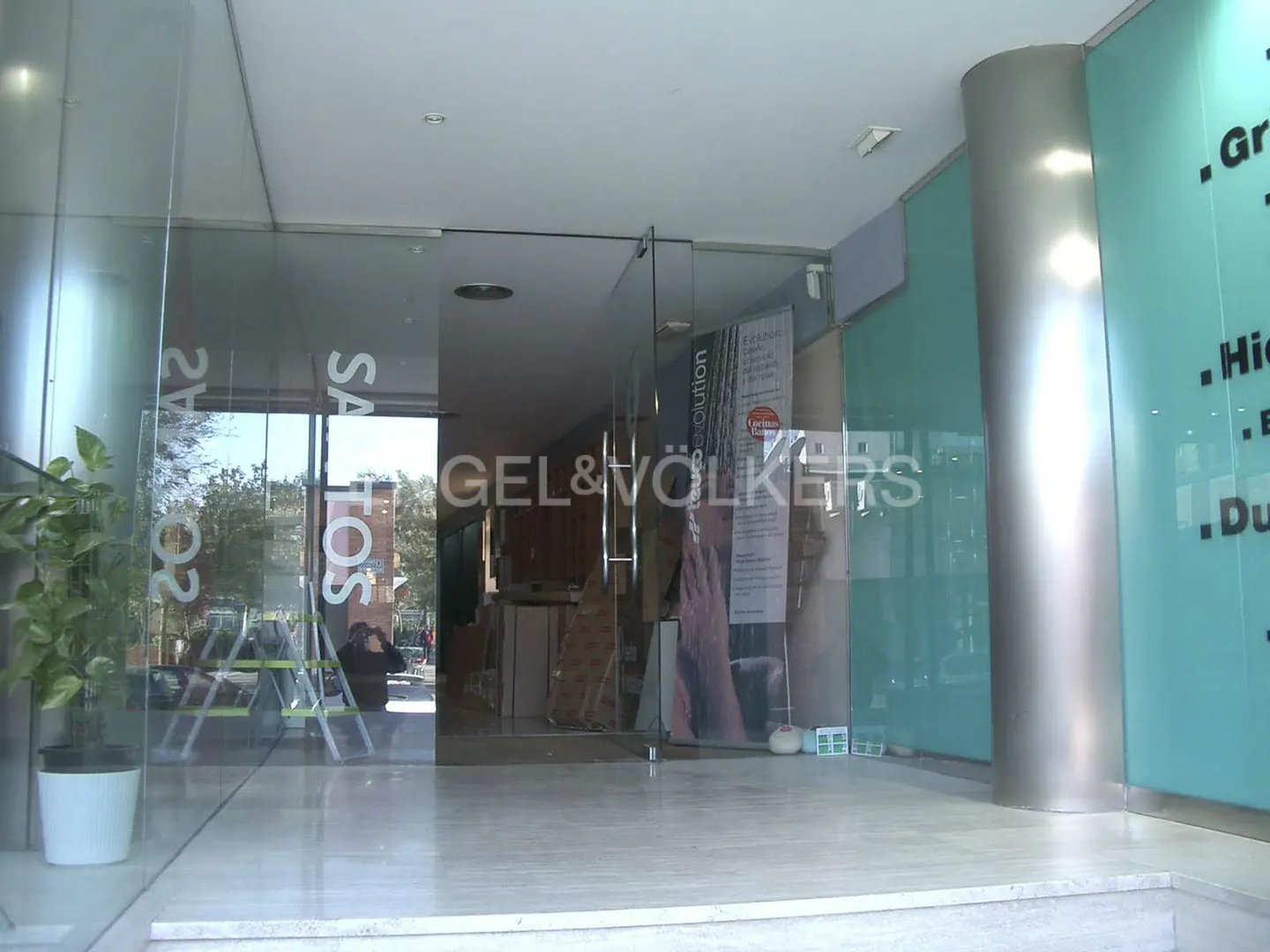 Exterior view of a Gel & Volkers real estate office with glass doors and windows. A potted plant sits near the entrance. The building has a modern design with turquoise accents.