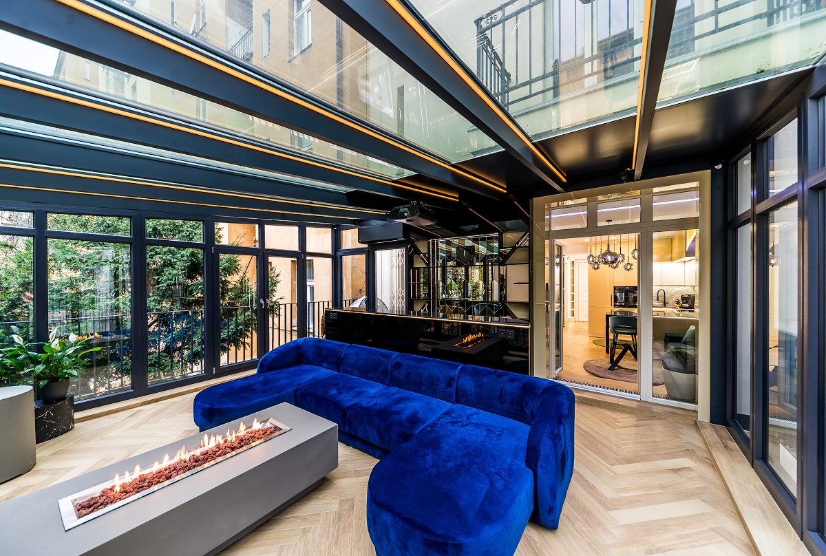Modern sunroom with a blue velvet sectional, linear fireplace, and glass ceiling, looking into a bright kitchen. [AI-generated text]