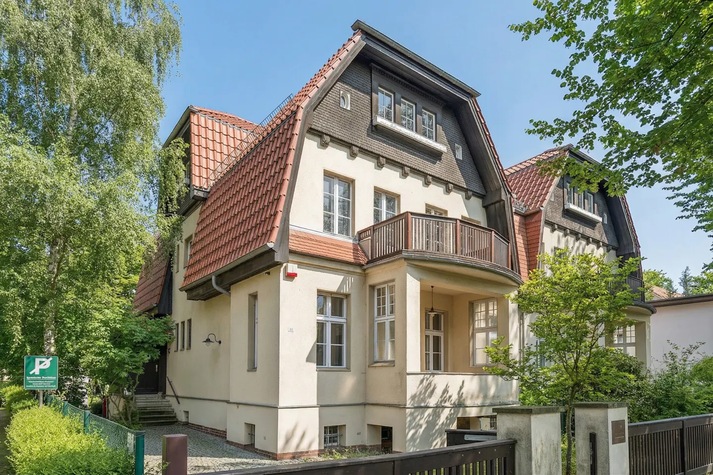 A three-story beige house with a red tile roof and a small balcony, surrounded by green trees.