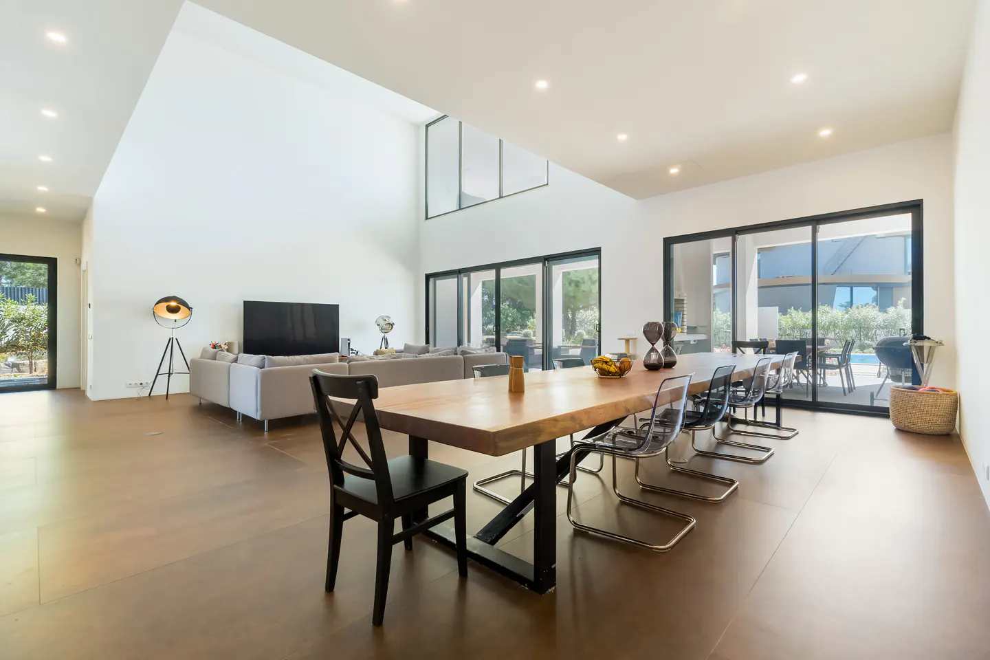 Bright, modern living space with a long wood table, clear chairs, and a gray sofa. Large windows offer a view of the outdoor pool area.