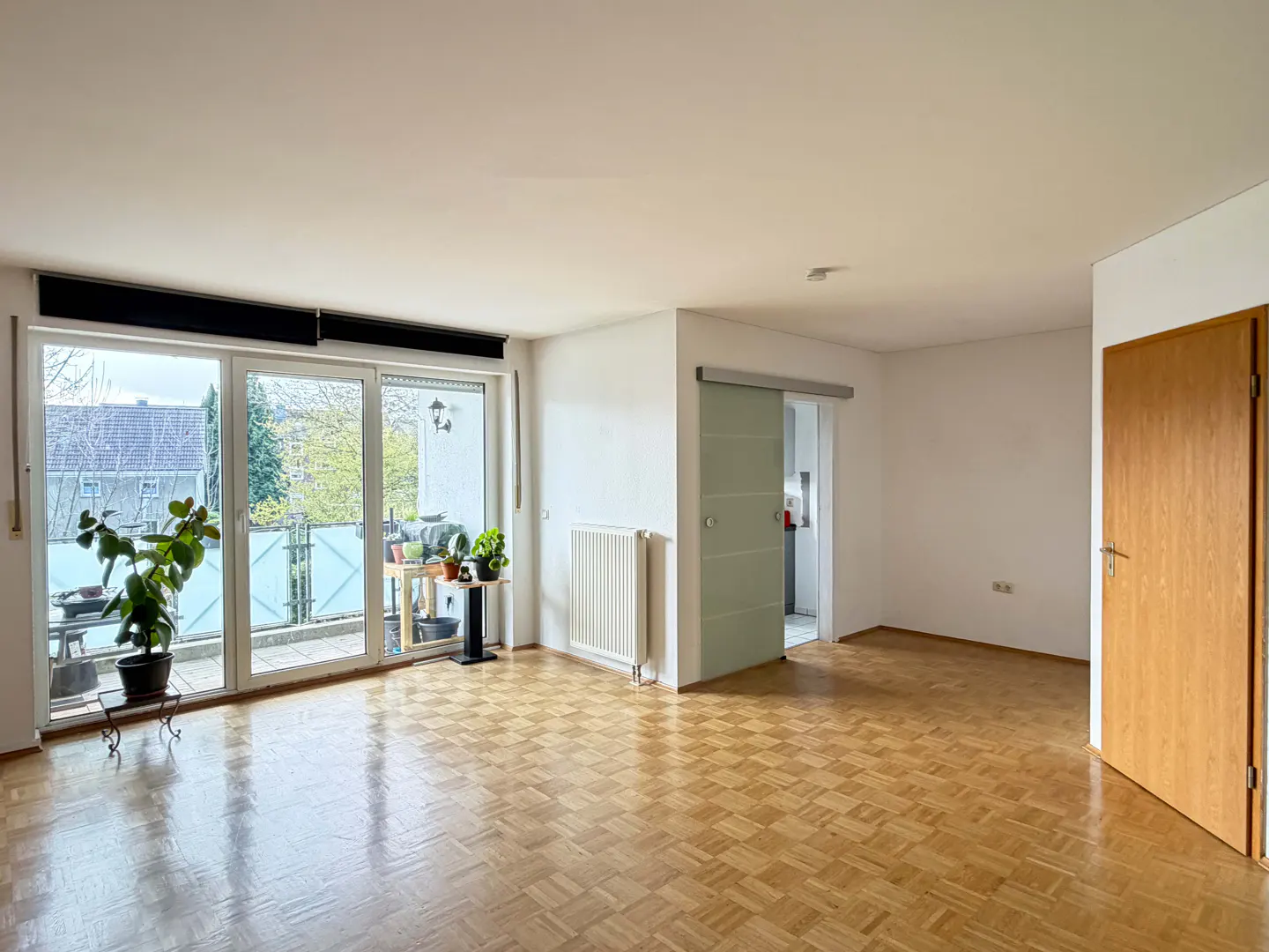 Bright, empty room with parquet floors, white walls, and a sliding glass door to a balcony with plants. A wooden door is on the right.
