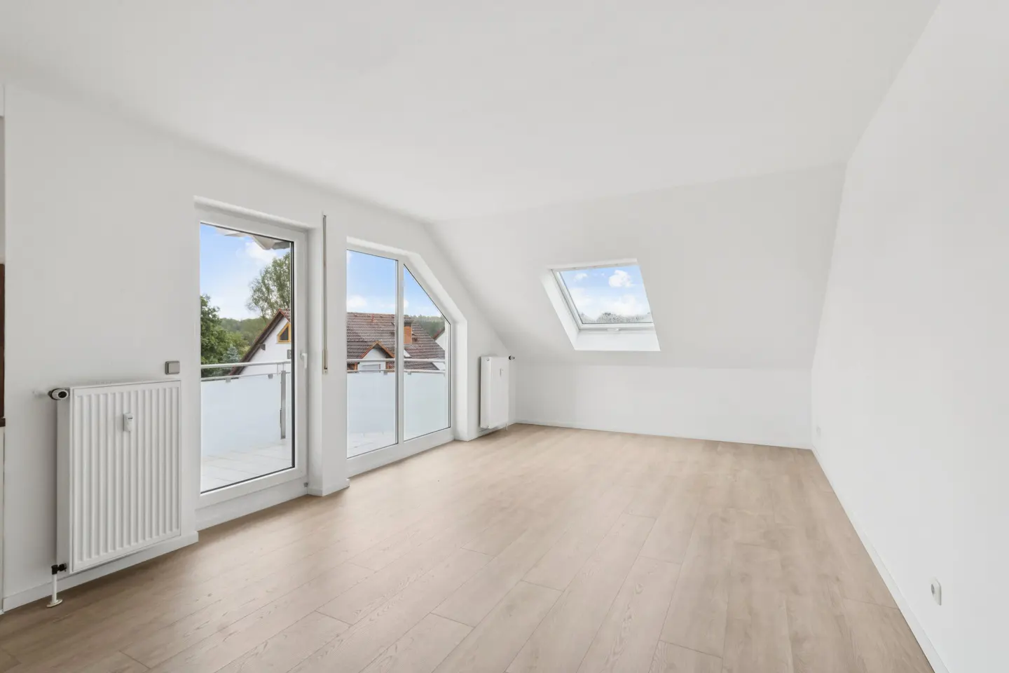 Bright, empty attic room with light wood floors, white walls, and a skylight. Balcony doors offer a view of trees and houses.