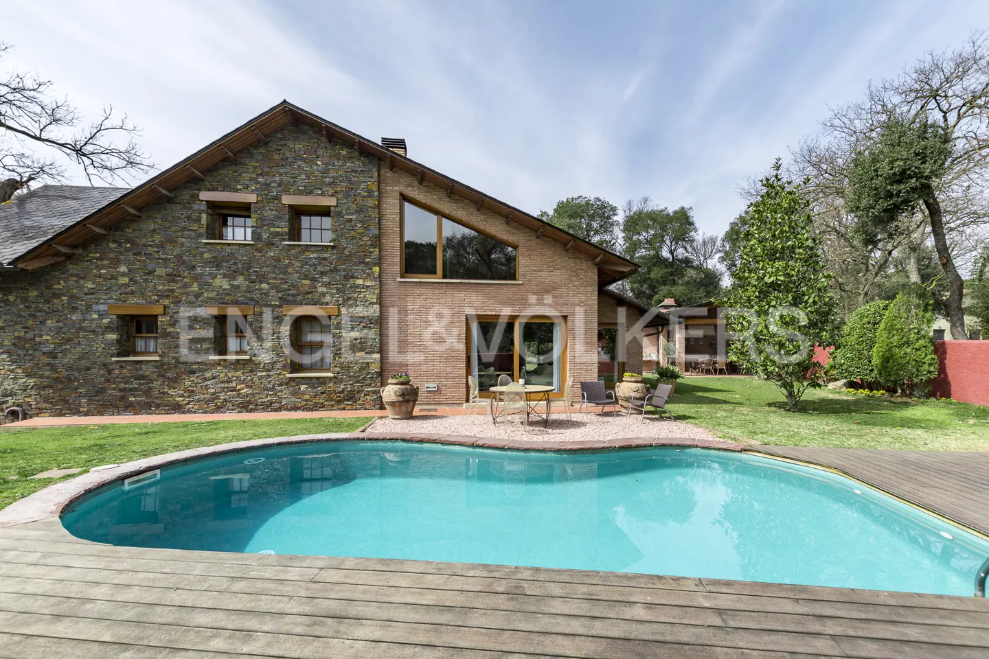 Exterior view of a stone and brick house with a blue swimming pool and wooden deck. A table and chairs sit on a gravel patio.