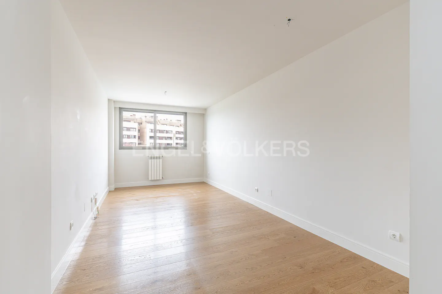 An empty room with light wood floors, white walls, and a window overlooking a city. A radiator is under the window.
