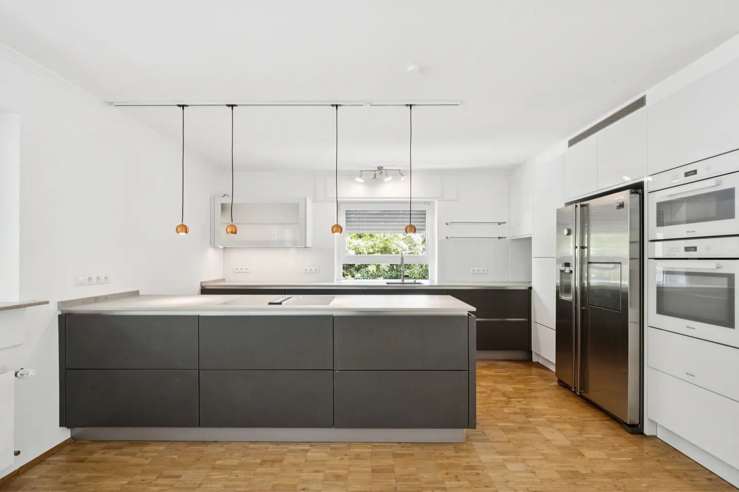 Bright kitchen with gray island, white cabinets, stainless steel fridge, and wood floors. Three pendant lights hang above the island.