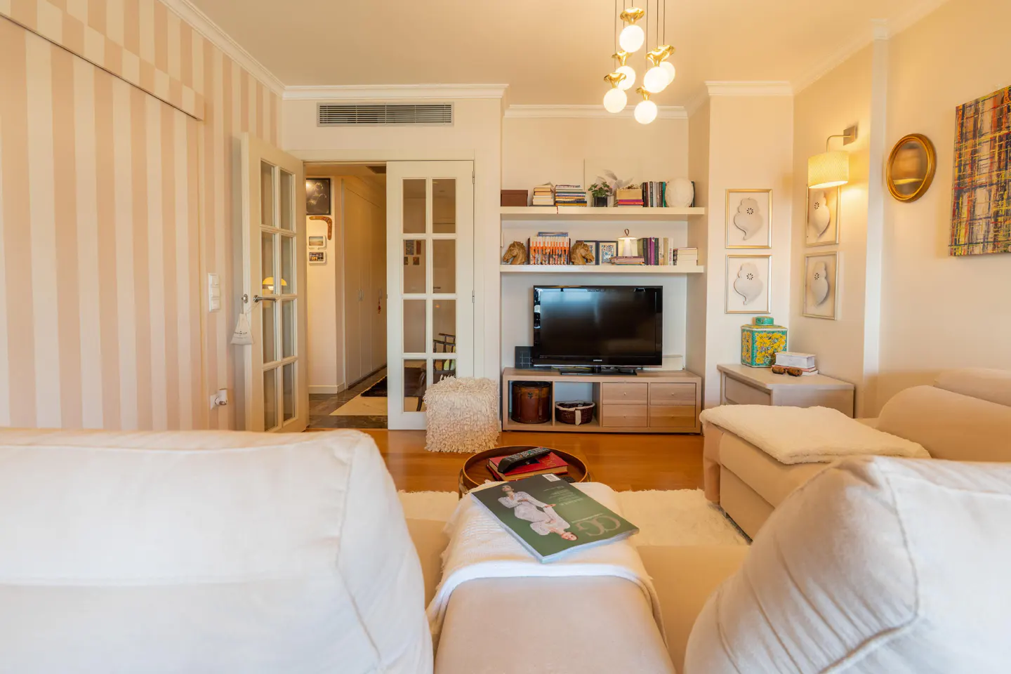 Living room with white sofas, a TV on a wooden stand, and a bookshelf. French doors lead to another room. Striped wallpaper.