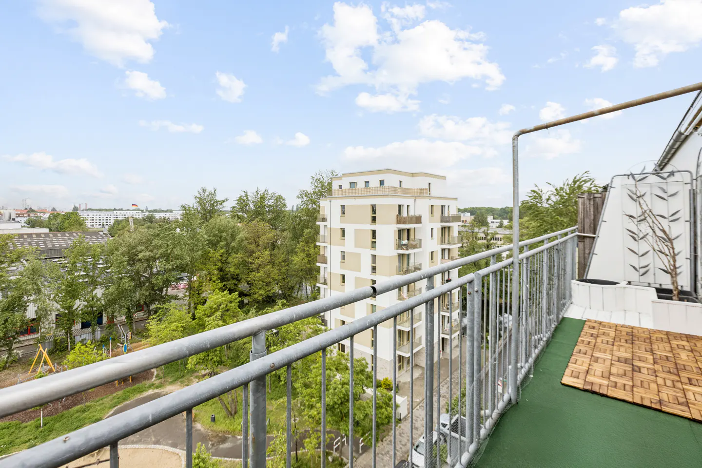 View from a balcony overlooking a green park and a modern multi-story apartment building under a blue sky with clouds.