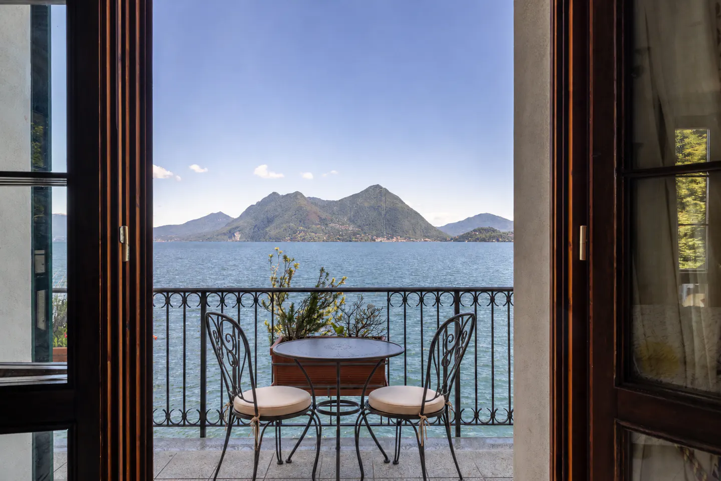 View from open doors to a balcony with a table and chairs overlooking a lake and mountains.
